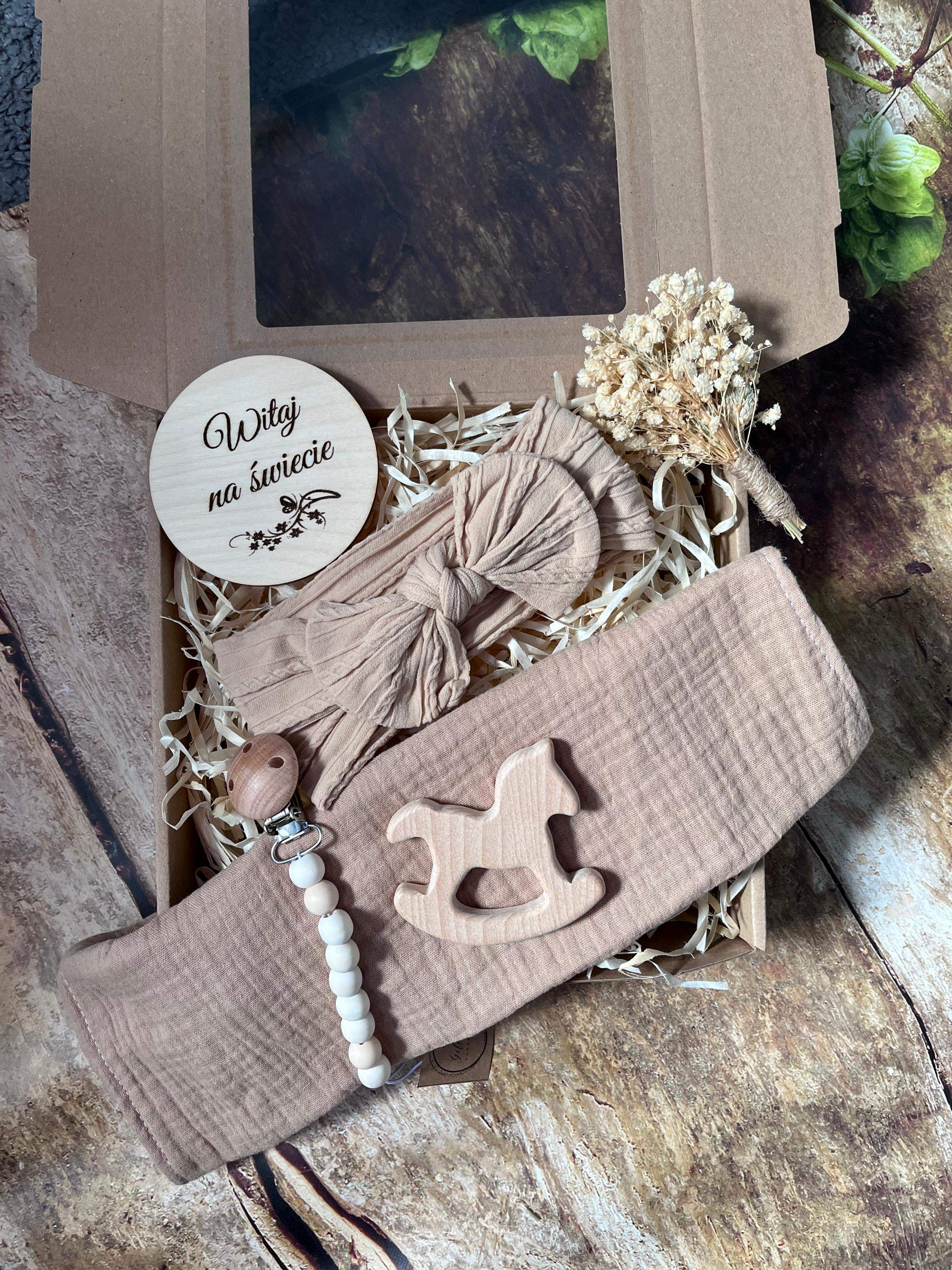 Tabliczka z grawerem wykonana z drewna, z napisem Witaj na świecie,  Zawieszka do smoczka ozdobiona drewnianymi i silikonowymi elementami, z możliwością personalizacji (np. imieniem dziecka). Muslinowa pieluszka – w odcieniu szarości, wykonana z miękkiego materiału, idealna dla noworodka. Gryzak w kształcie słonika – wykonany z drewna, bezpieczny i przyjazny dla dziecka. Szczotka dla niemowlaka – z naturalnym, miękkim włosiem i drewnianą rączką. Skarpetki niemowlęce – w kolorze szarym, miękkie i wygodne dla malutkich stópek.